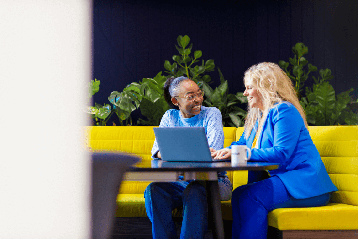 Two women working together infront of a laptop at a table sat on a large yellow sofa. One woman is wearing a light blue jumper and jeans. The other is in a blue suit.
