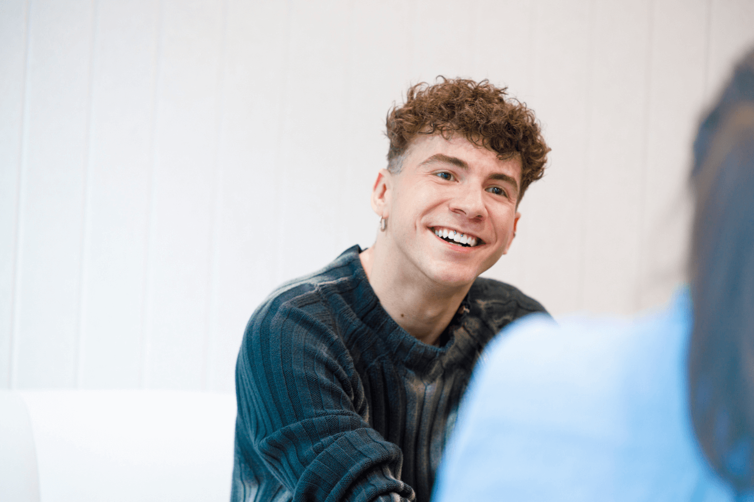 A man with black curly hair and a black jumper smiling at the camera. 