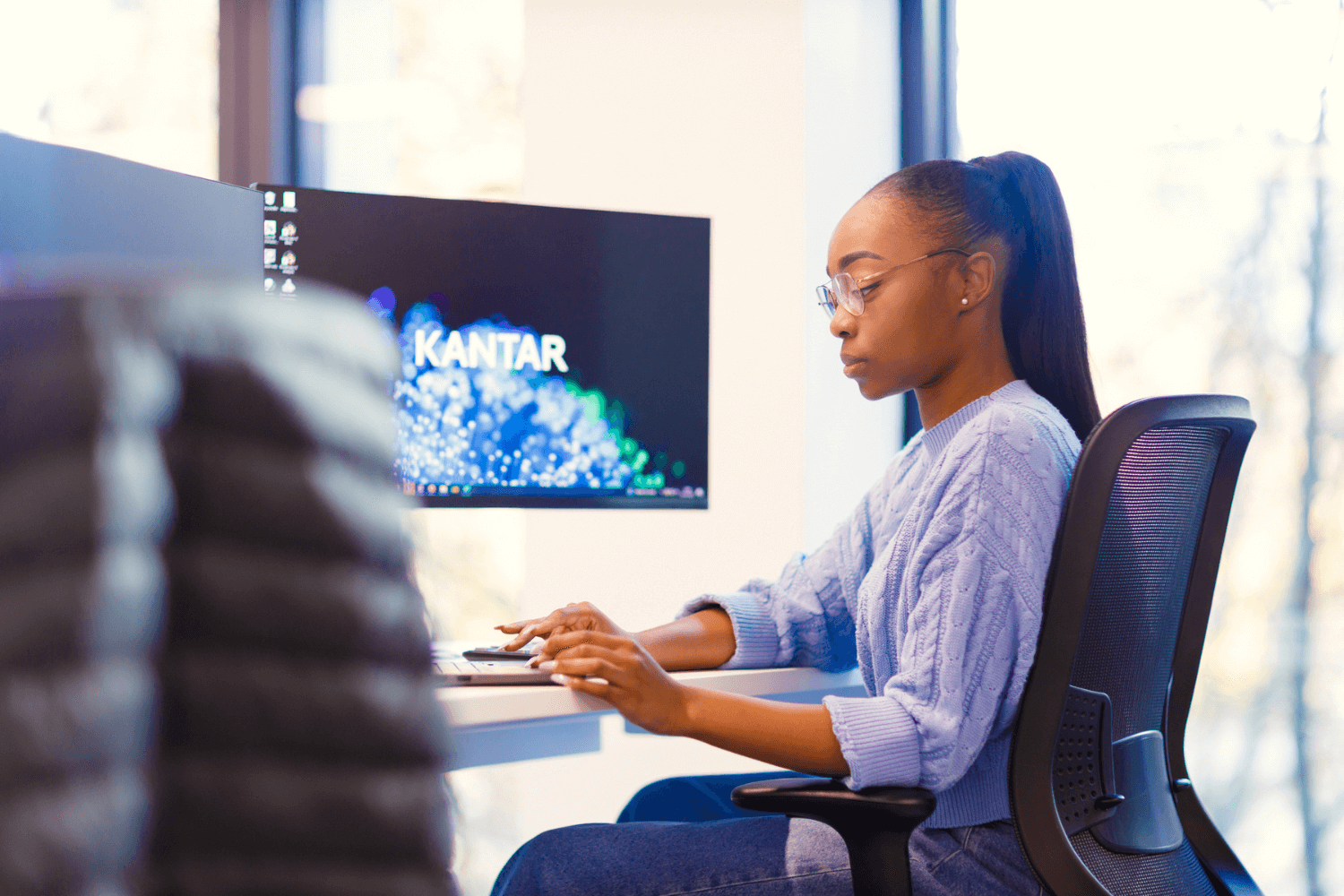 A woman working at her desk. She is wearing a light blue jumper and glasses, with long hair in a ponytail. 