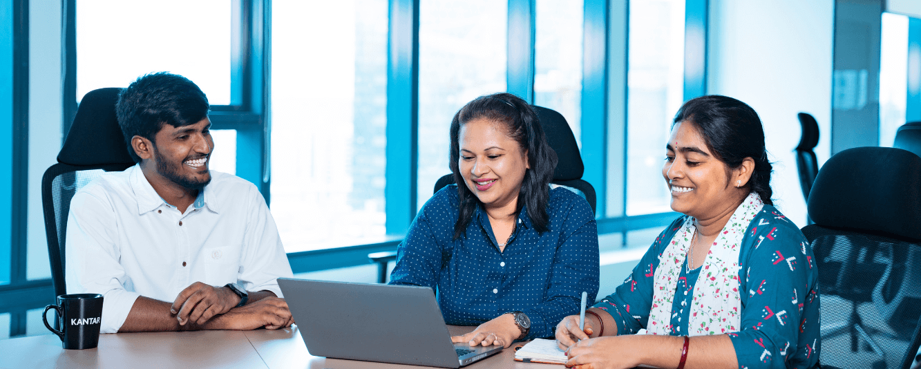 Three Indian colleagues are working together. From left to right. A man with a short brown beard is smiling across at two female colleagues. He is wearing a white shirt. In the middle, a female colleague is working at a silver laptop and has dark hair to just past her shoulders. She is wearing a blue shirt. Finally there is a second woman making notes in her notebook. She is wearing a white patterned scarf and blue and red patterned elbow length top. Her hair is in a braid.