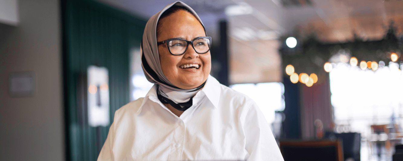 A woman in a white shirt smiles off to the right. She is wearing a headscarf, which is a mix of light and dark colours. She is wearing glasses.