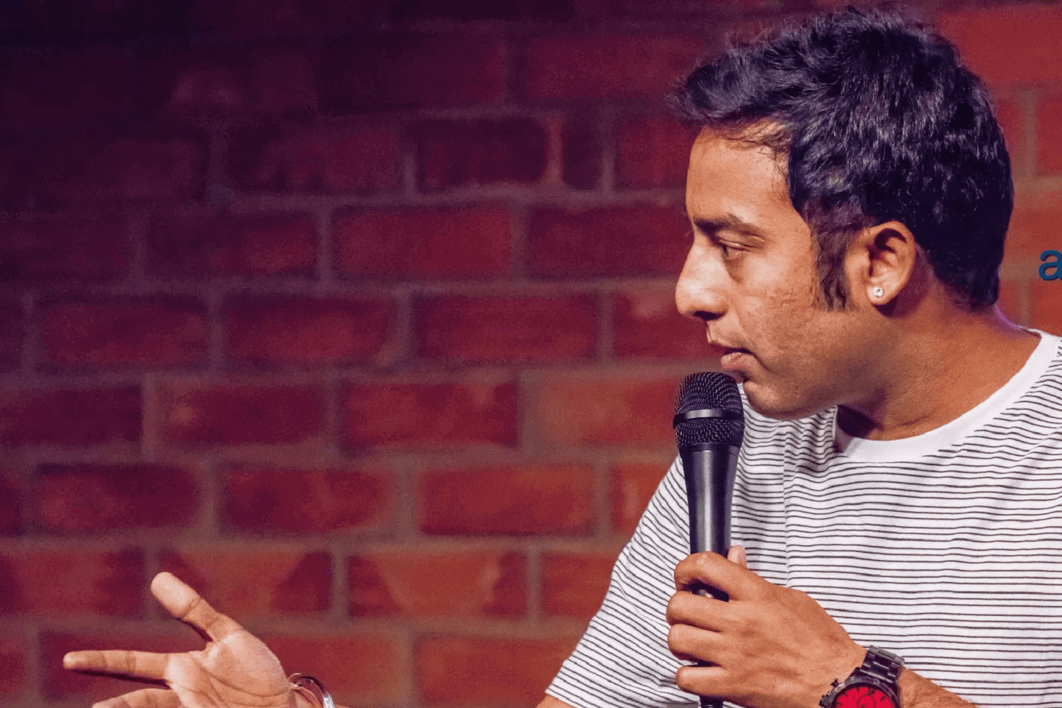 A man on stage. He is a grey and light horizontally striped T-shirt and holding a microphone. 