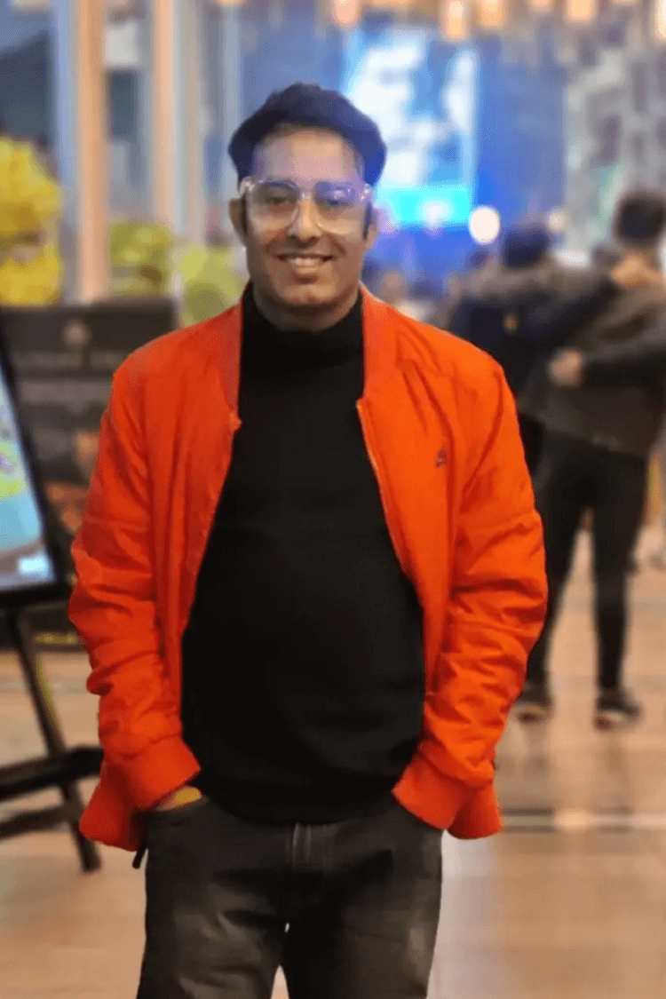 A man in an orange jacket and a black polo, smiling directly at the camera. He is outside in a cafe type scene. 