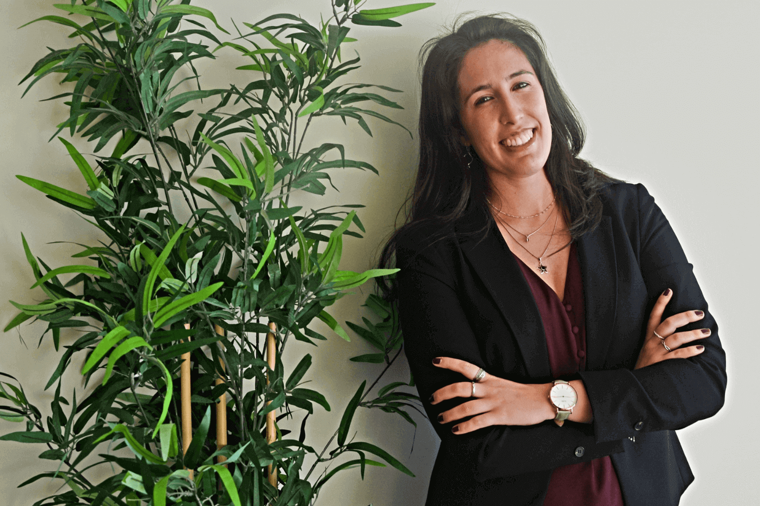 A woman leaning next to a plant. She has long brown hair and is wearing a black jacket and red dress. 