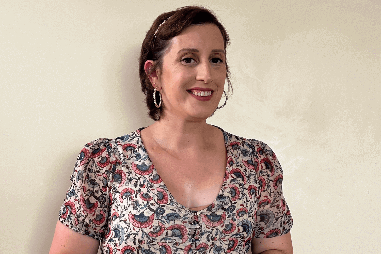 A woman with short brown hair and gold hooped earrings. She is wearing a multicoloured, patterned top. 