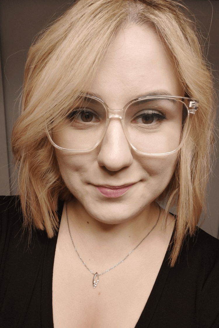 A woman with a black top and glasses with transparent frames. She has shoulder length blonder hair. 