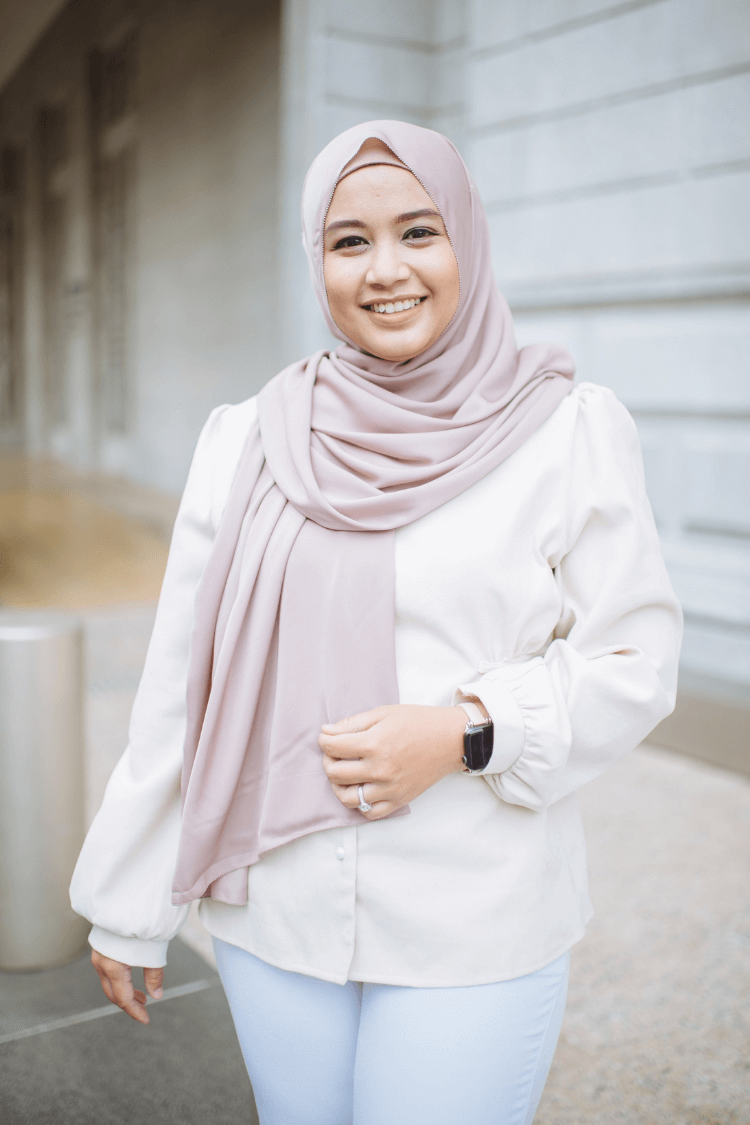 A woman with a pink head covering and a long sleeved white top, with jeans. 