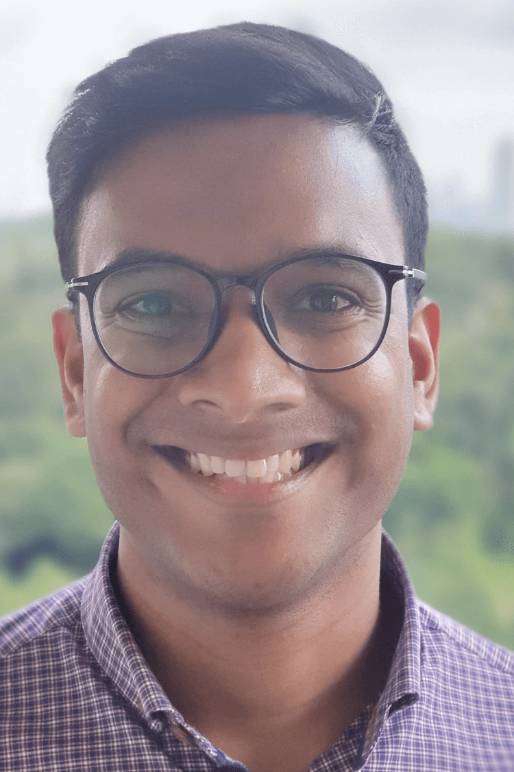 A young man with round, black rimmed glasses smiling at the camera. He is wearing a checked shirt and he has short brown hair. 