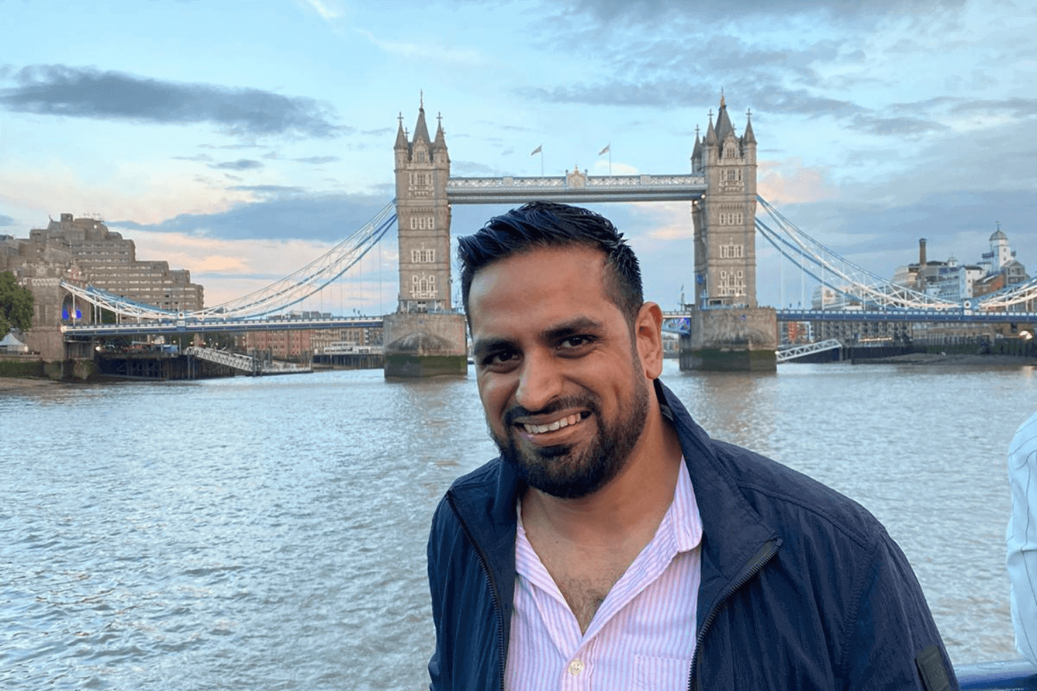 A man with a blue jacket and light shirt. Tower Bridge is behind him. 