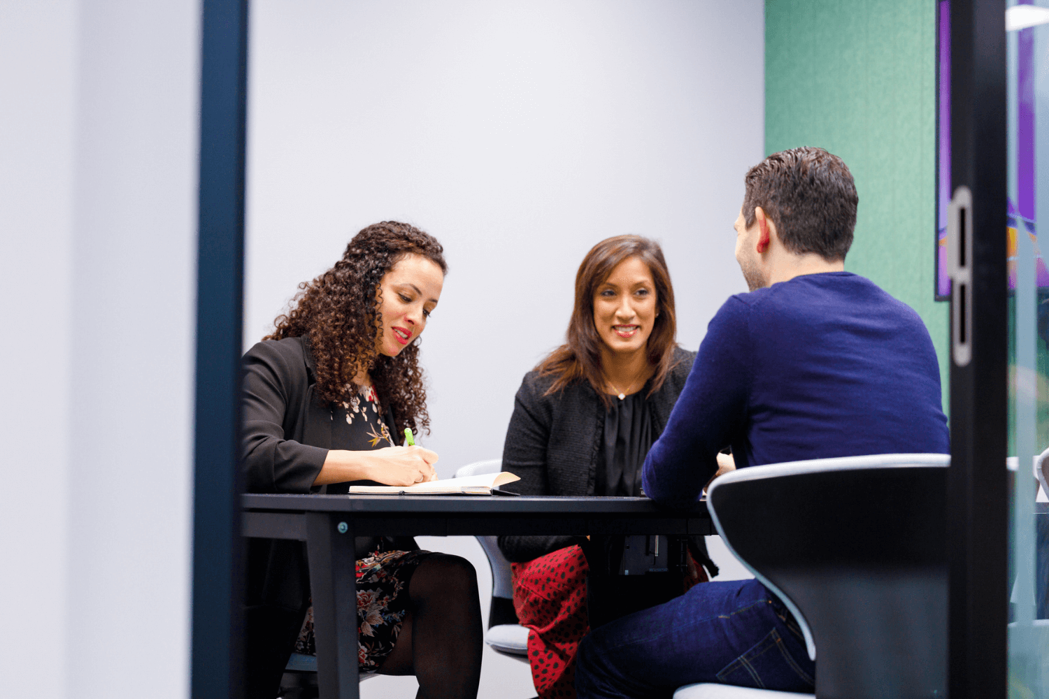 Três colegas em uma reunião. A sala de reunião é verde, com uma mesa no meio. Há duas mulheres voltadas para a frente e um homem de costas para a câmera.
