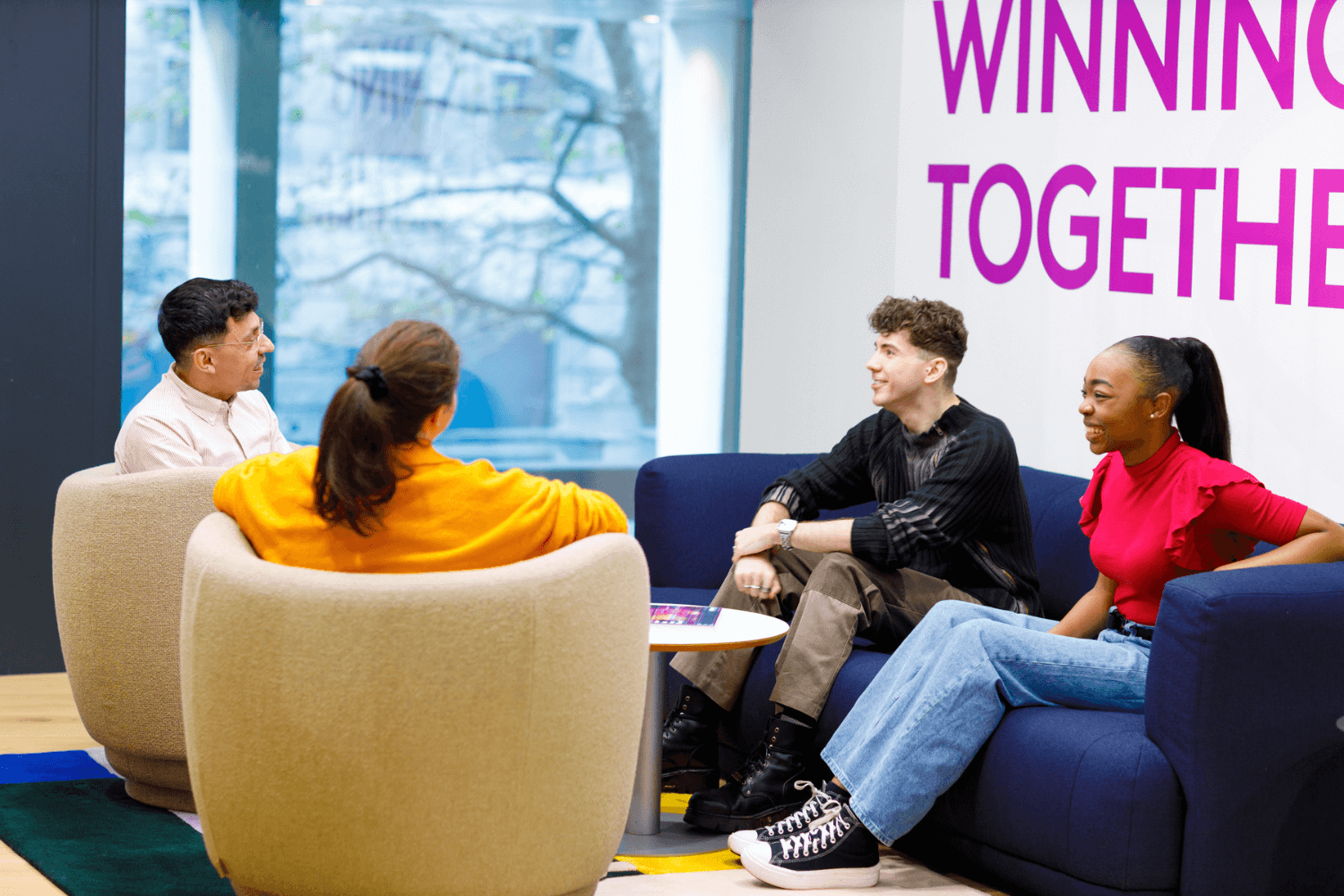 Eine Gruppe von vier Personen sitzt auf dem Sofa und unterhält sich, zwei Männer und zwei Frauen. An der Wand hinter ihnen steht "Winning Together".