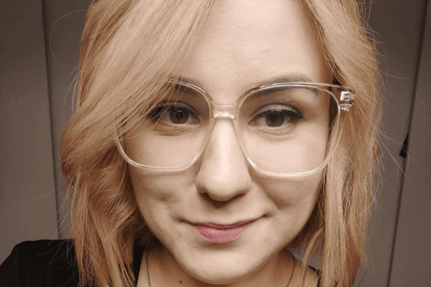 Eine Frau mit einem schwarzen Oberteil und einer Brille mit durchsichtigem Gestell. Sie hat schulterlanges, blondes Haar. 