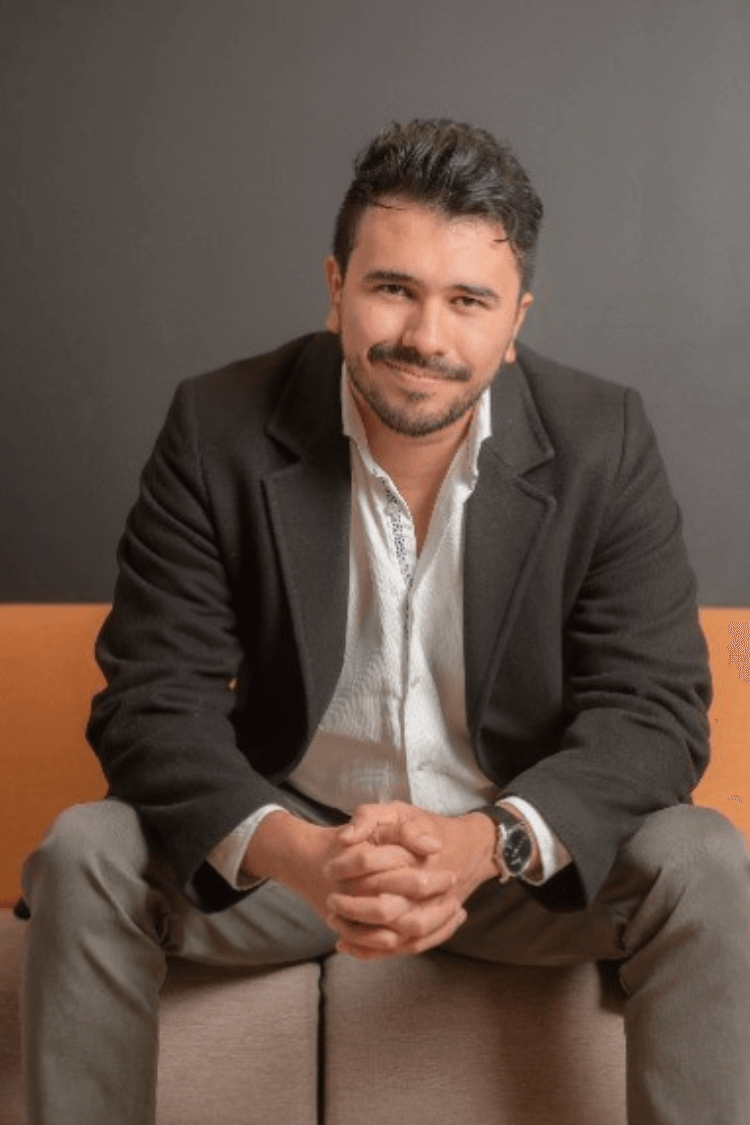 Ein Mann sitzt auf einem orange-braunen Sofa. Er trägt eine graue Chinohose, ein weißes Hemd und eine graue Jacke. 