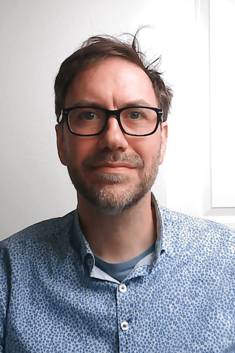 Ein Mann mit einer schwarz umrandeten Brille und einem blauen Hemd schaut in die Kamera. Er hat kurzes braunes Haar. 