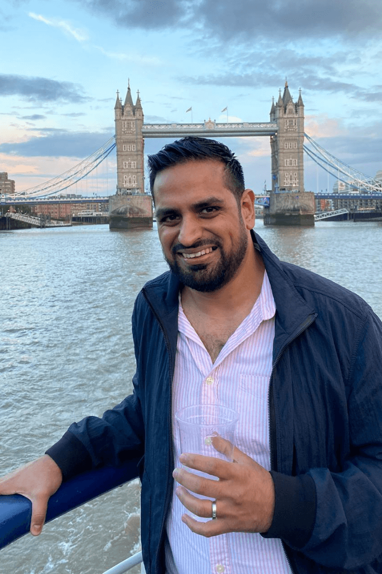 Ein Mann mit einer blauen Jacke und einem hellen Hemd. Die Tower Bridge liegt hinter ihm. 