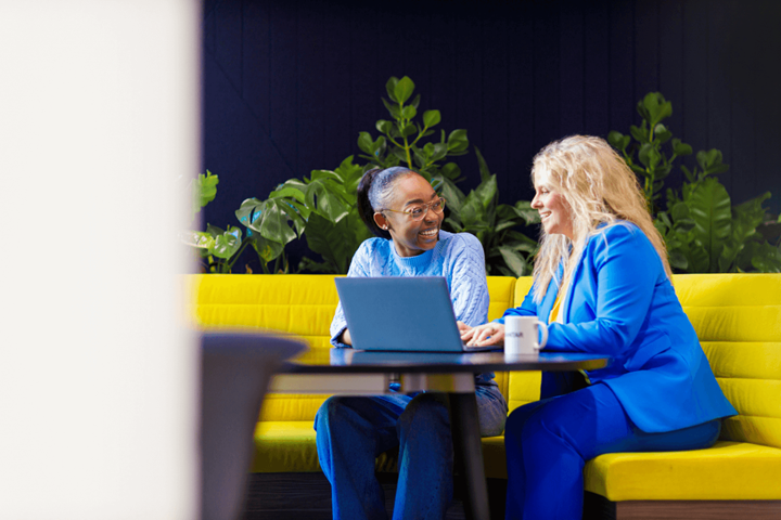 Two women working together infront of a laptop at a table sat on a large yellow sofa. One woman is wearing a light blue jumper and jeans. The other is in a blue suit.