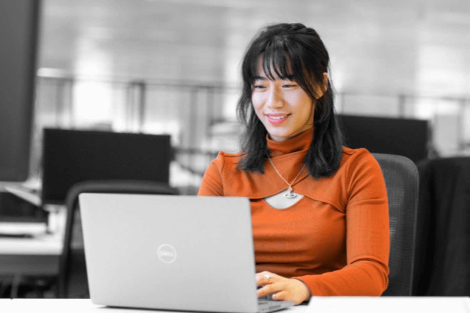 Image en noir et blanc, où seuls les vêtements et le teint de la peau sont en couleur. Une jeune femme vêtue d'un pull-over orange travaille sur un ordinateur portable.