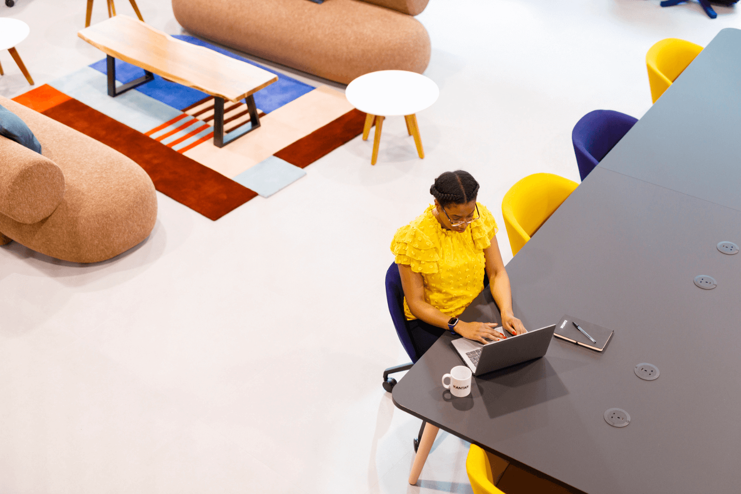 Une femme travaillant sur un ordinateur portable à une longue table. Elle porte un haut jaune. La photo est prise du dessus.