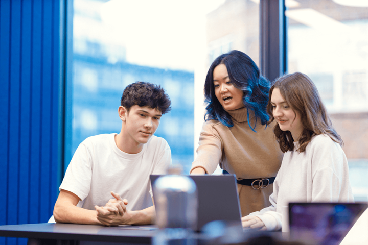 Een groep van drie mensen rond een laptop. Een vrouw in een bruine trui met lang bruin haar en blauwe highlights legt iets uit aan twee jongere collega's. De ene is een man en de andere een vrouw. De ene collega is een man en de andere een vrouw; beiden dragen een wit topje.