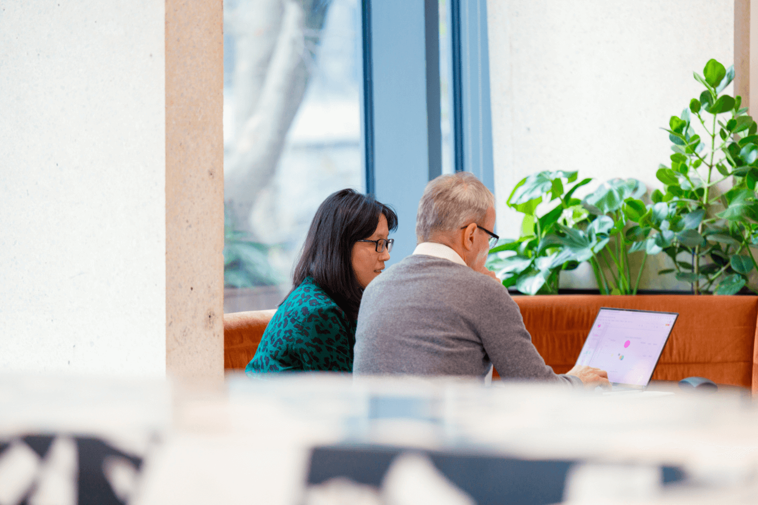 Twee collega's werken samen vanaf een laptop. Zij heeft schouderlang donker haar en draagt een bril en een groene jurk. Hij draagt een middengrijze trui en een wit overhemd. 