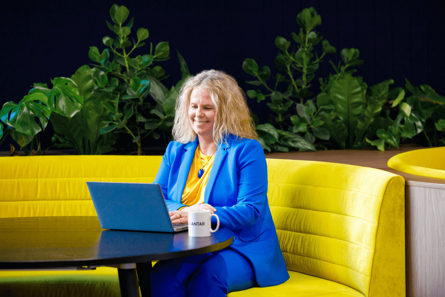 Een vrouw met lang krullend blond haar zat op een gele bank aan een laptop te werken.
