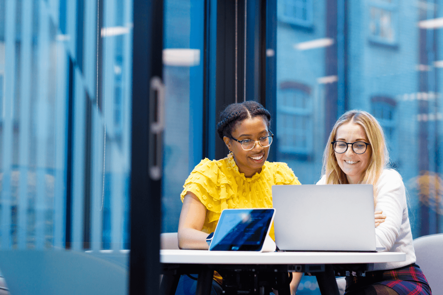 Twee vrouwen werken samen aan een laptop in een vergaderruimte. De een draagt een gele blouse, de ander een witte trui.