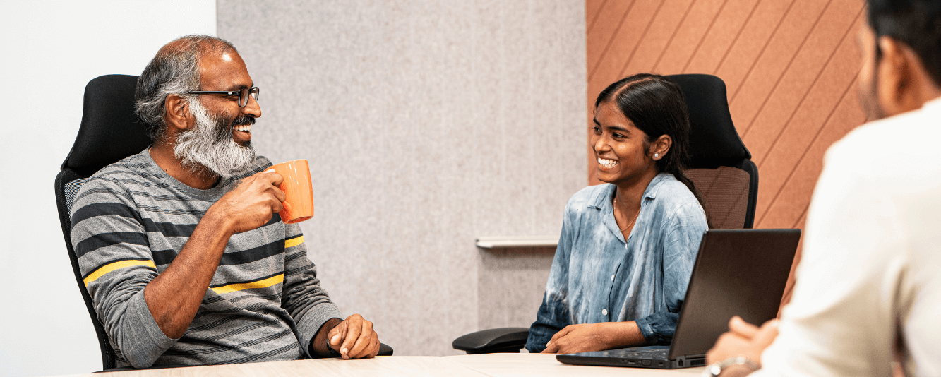Twee collega's praten samen. Links is een oudere heer. Hij is Indiaas, heeft een grijze baard en draagt een grijze trui met zwarte en gele strepen. Hij drinkt uit een oranje mok. Hij praat met een jongere vrouwelijke collega. Zij draagt een blauwe blouse en heeft haar haar naar achteren gebonden.