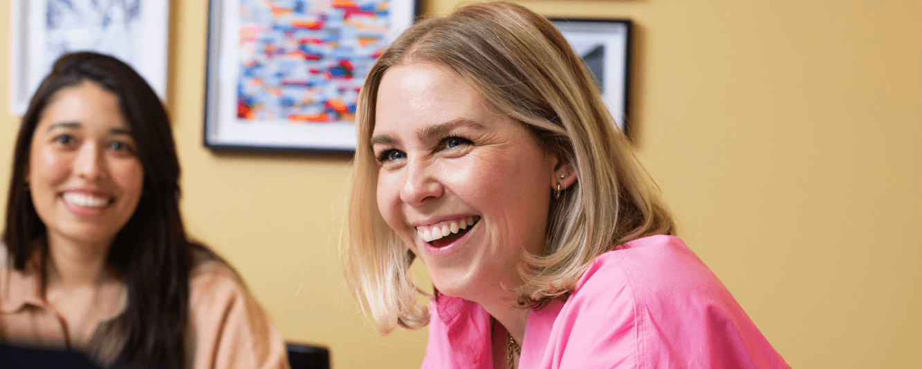 Een vrouw met kort blond haar en een roze shirt lacht naar de camera. Ze zit in een kamer die felgeel is geschilderd. Achter haar is een andere vrouwelijke collega, enigszins onscherp. Ze heeft lang bruin haar en draagt een bruin topje.