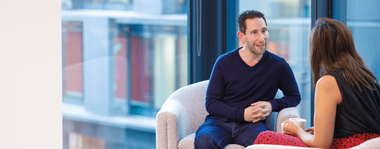 Een man die een gesprek voert met een vrouwelijke collega, terwijl hij op comfortabele stoelen zit.