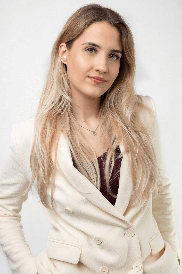 Een vrouw met lang blond haar, die een witte jas met knopen draagt, met daaronder een rood topje. 