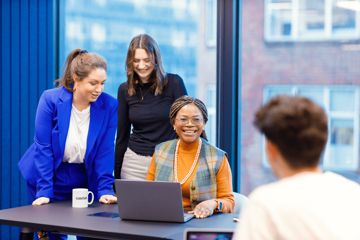 四位同事在一起开会。其中有三位女性和一位男性。其中一位女士正用笔记本电脑工作，对着镜头微笑，其他人则站在她周围，看着屏幕。 